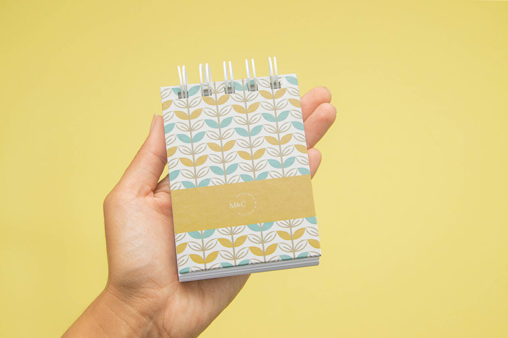 Product Photography for Minico Person holding a yellow, white, and blue notebook with leaf abstract design in their palm on yellow background, photographed by Atlanta product photographer Natalia Tamariz, showcasing stylish stationery for small brands.