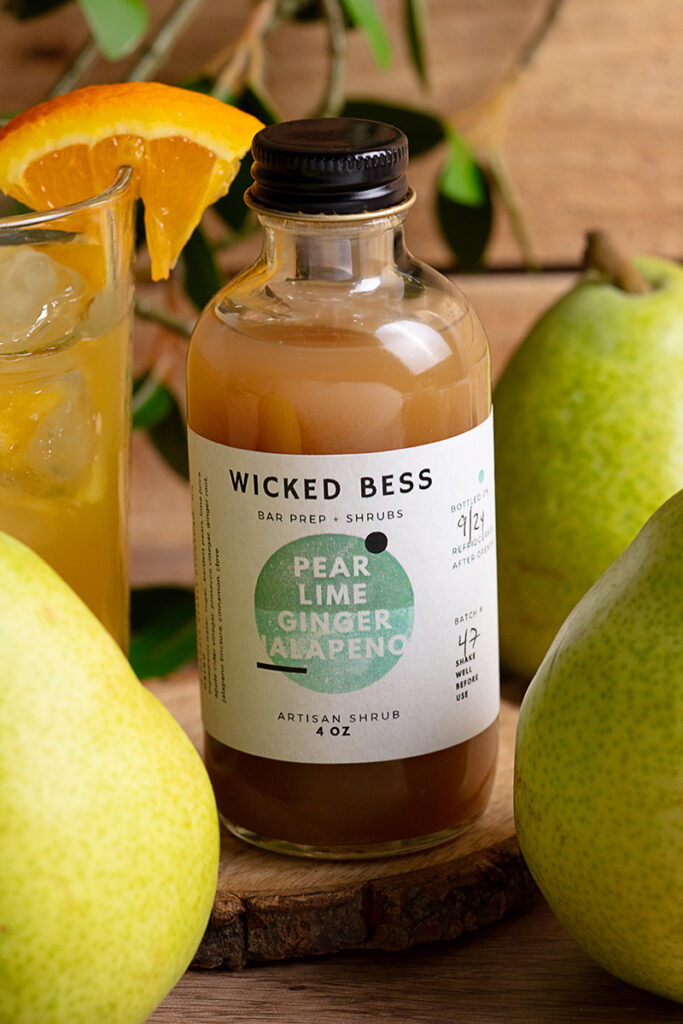 Wicked Bess product photography Wicked Bess juice bottle with oranges, a glass of juice, and green background, photographed by Atlanta product photographer Natalia Tamariz, showcasing fresh and vibrant beverage products for small brands.