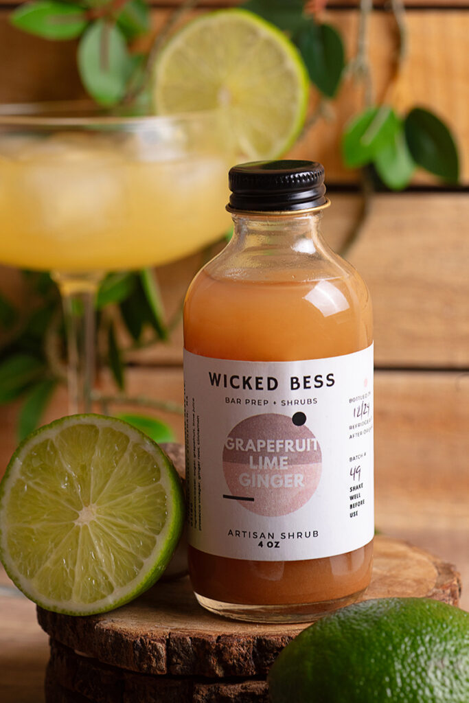 Wicked Bess product photography Wicked Bess juice bottle with oranges, a glass of juice, and green background, photographed by Atlanta product photographer Natalia Tamariz, showcasing fresh and vibrant beverage products for small brands.