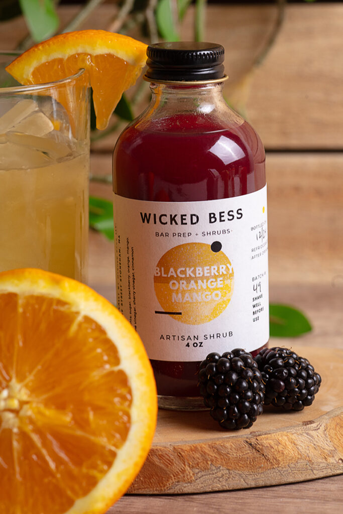 Wicked Bess product photography Wicked Bess juice bottle with oranges, a glass of juice, and green background, photographed by Atlanta product photographer Natalia Tamariz, showcasing fresh and vibrant beverage products for small brands.